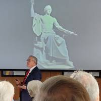 A man speaks in front of a buff George Washington statue